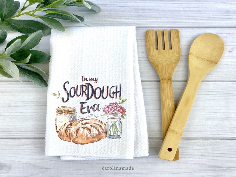 This white waffle weave towel says In my sourdough era and shows a jar of dough, bread, and a jar of flowers. The perfect kitchen towel for bakers.