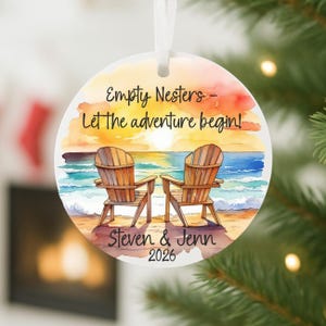 May include: Round Christmas ornament with a beach scene featuring two wooden Adirondack chairs on a sandy shore. The ornament has the text "Empty Nesters - Let the adventure begin!" and the names "Steven & Jenn" with the year "2026".