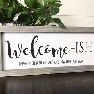 May include: A gray wooden framed sign with the text "Welcome-ish" and the subtext "Depends on who you are and how long you stay".