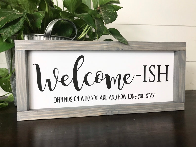 May include: A gray wooden framed sign with the text "Welcome-ish" and the subtext "Depends on who you are and how long you stay".