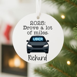 May include: A white, round Christmas ornament with a ribbon for hanging. The ornament features the text "2025: Drove a lot of miles." and a black car graphic with the word "UBER" in blue. The name "Richard" is written below the car.