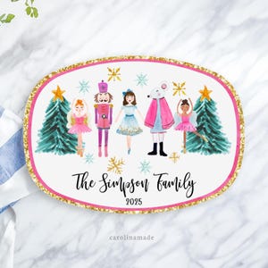 May include: An oval-shaped, white decorative plaque with a pink border and gold glitter trim. The plaque features a watercolor illustration of characters from The Nutcracker ballet, with text that reads "The Simpson Family 2025".