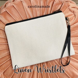 Puede incluir: Una cartera de mu&ntilde;eca de lino blanco con una cremallera negra y una correa de mu&ntilde;eca negra. La cartera de mu&ntilde;eca est&aacute; sobre un fondo rosa. El texto "Linen Wristlets" est&aacute; escrito debajo de la cartera de mu&ntilde;eca.
