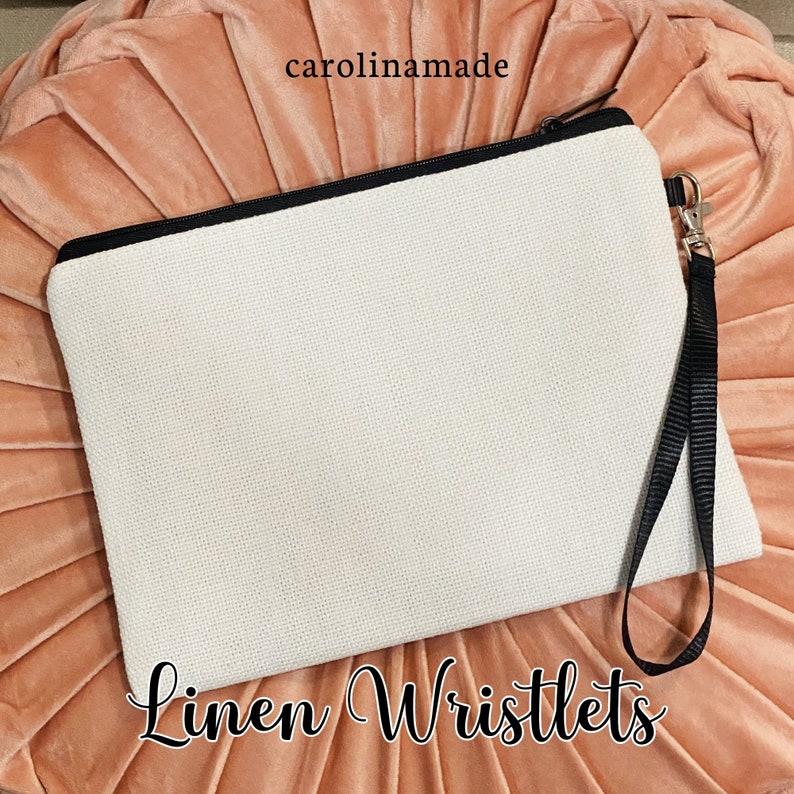 Puede incluir: Una cartera de mu&ntilde;eca de lino blanco con una cremallera negra y una correa de mu&ntilde;eca negra. La cartera de mu&ntilde;eca est&aacute; sobre un fondo rosa. El texto "Linen Wristlets" est&aacute; escrito debajo de la cartera de mu&ntilde;eca.