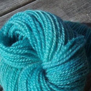 以下が含まれることがあります： 鮮やかなターコイズブルーの糸の玉が、きつく巻かれた状態。糸は柔らかくふわふわで、色は鮮やかなターコイズブルーです。