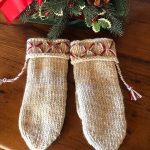 May include: A pair of beige knitted mittens with red, white, and blue trim. The mittens are on a wooden surface.