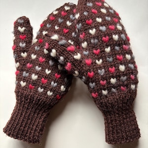 May include: A pair of brown knitted mittens with a heart pattern in red, white, and gray. The mittens have a ribbed cuff and a textured appearance, perfect for cold weather. The heart pattern is evenly distributed across the surface.