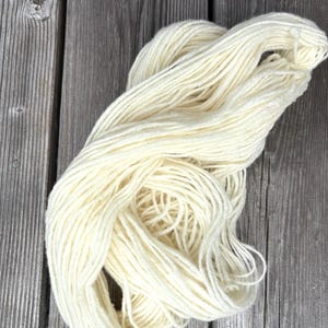 以下が含まれることがあります： 使い古された木材の表面に、オフホワイトの糸の束が置かれています。糸はきつく巻かれており、糸の束と柔らかい質感が見えます。ニュートラルな色と天然繊維は、手芸品やテキスタイル製品を示唆しています。