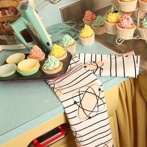 May include: A white tea towel with black stripes and pink atomic symbols. The tea towel is folded and placed on a yellow countertop with a teal blue countertop behind it. There are cupcakes on a tiered stand and a teal blue hand mixer on the countertop.