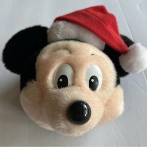 May include: A plush Mickey Mouse head ornament featuring a soft beige face, large black ears, and a bright red Santa hat with a white pom-pom. The character has prominent white eyes with black pupils and a small, round, shiny black nose.