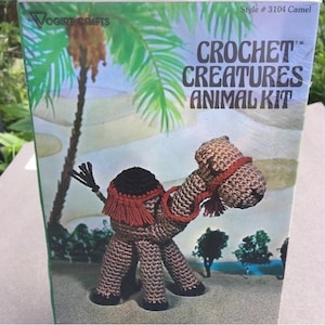 May include: A Vogart Crafts Crochet Creatures Animal Kit box featuring a brown crocheted camel with a red saddle and harness. The packaging displays the text Style # 3104 Camel and Crochet Creatures Animal Kit against a desert background.