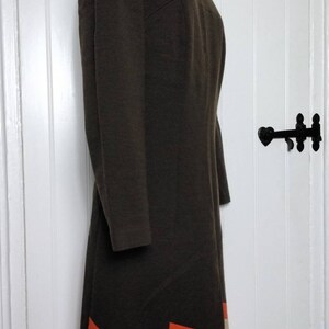 May include: A brown dress with a simple, round neckline and long sleeves. The hem of the dress features a geometric pattern of orange and cream coloured stripes.