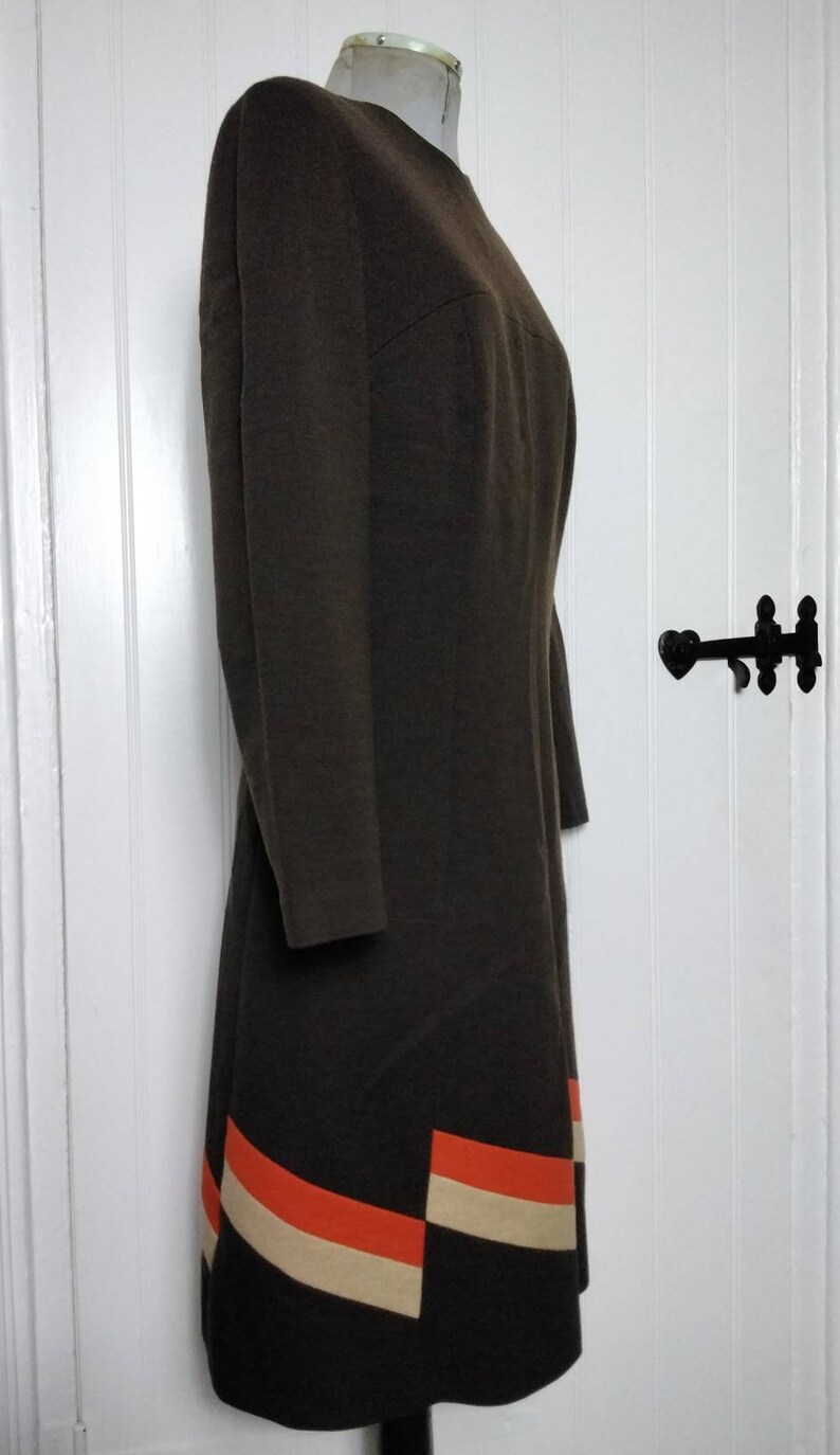 May include: A brown dress with a simple, round neckline and long sleeves. The hem of the dress features a geometric pattern of orange and cream coloured stripes.