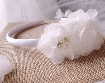 May include: A white headband featuring fabric flowers and pearl and rhinestone embellishments. The flowers have a layered, delicate design with a soft, sheer texture. The headband is displayed on a neutral surface.