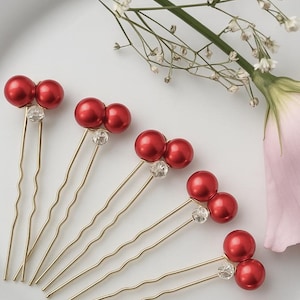 Op de afbeelding: Vijf goudkleurige haarspelden, elk met twee rode imitatieparels en een helder kristallen accent. De spelden zijn gerangschikt op een wit oppervlak, met een takje witte bloemen en een roze bloemblad op de achtergrond.