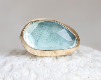 May include: A gold ring featuring a large, light blue gemstone with a faceted cut. The ring's setting has a hammered texture. The ring is displayed on a white, textured surface, creating a contrast of colors and textures.