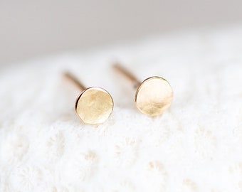 May include: A pair of small, round, gold stud earrings. The earrings are simple and minimalist, and they have a slightly hammered texture.
