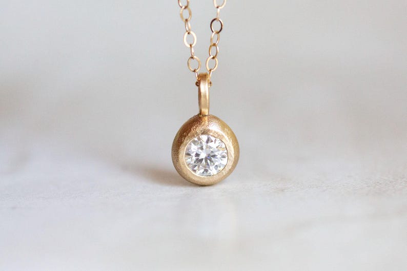 May include: A gold pendant necklace with a round, textured setting that holds a clear, round diamond.