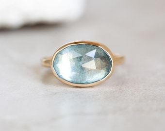 May include: A gold ring featuring a large, oval-shaped, light blue gemstone. The gemstone has a faceted surface, catching the light. The ring is set on a simple, thin gold band, showcasing the stone's color and shape.