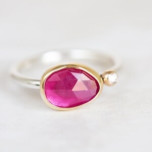 May include: A silver ring with a pink gemstone set in a gold bezel. A small diamond is set in the band.