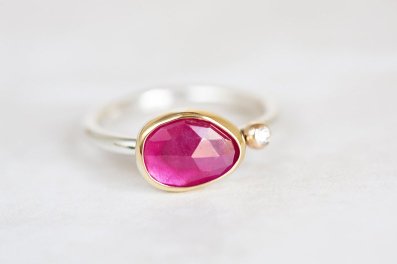 May include: A silver ring with a pink gemstone set in a gold bezel. A small diamond is set in the band.