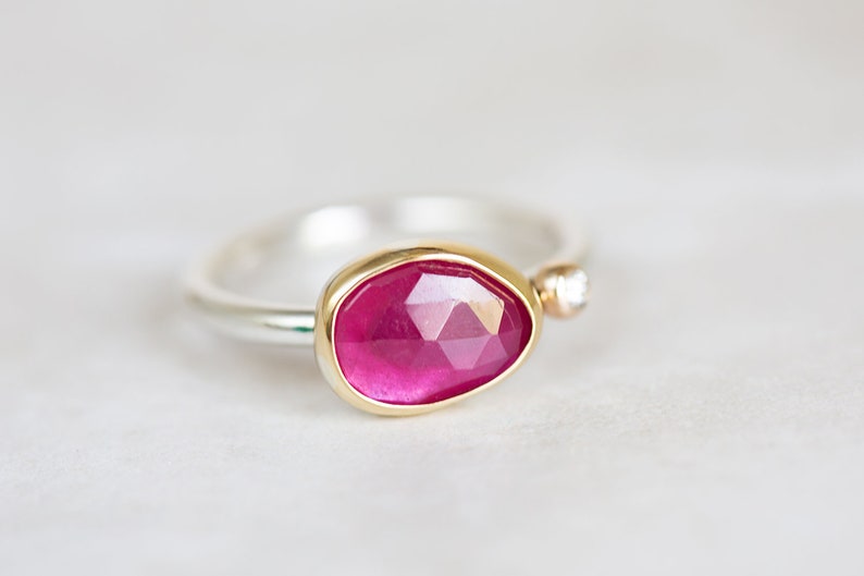 May include: A silver ring with a pink gemstone set in a gold bezel. A small diamond is set in the band.