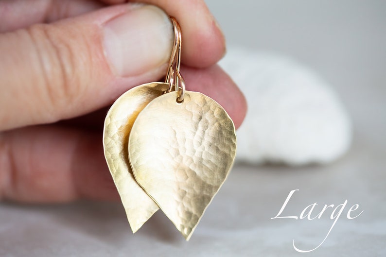 May include: Gold leaf earrings with a hammered texture. The earrings are large and have a teardrop shape. The word "Large" is written in cursive below the earrings.