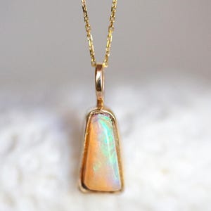 May include: A gold pendant necklace featuring a trapezoidal-shaped opal gemstone with iridescent flashes of green and orange. The opal is set in a smooth gold bezel frame and hangs from a delicate gold chain, showcasing a polished metallic finish.