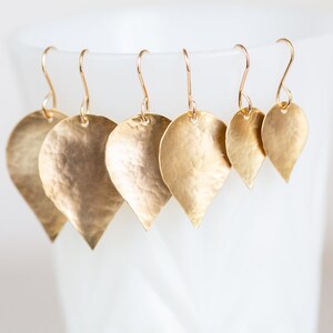 May include: A set of five gold-toned leaf-shaped earrings with hammered textures. The earrings are arranged in descending size order, with the largest earring at the top and the smallest earring at the bottom.
