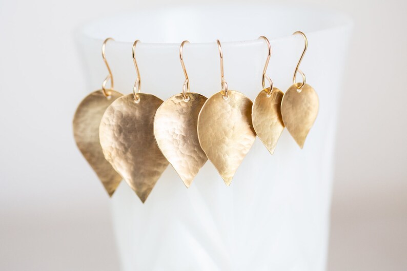 May include: A set of five gold-toned leaf-shaped earrings with hammered textures. The earrings are arranged in descending size order, with the largest earring at the top and the smallest earring at the bottom.