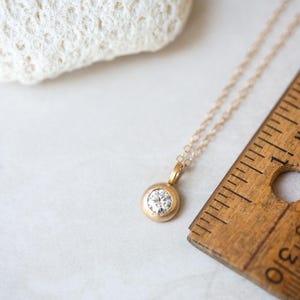 May include: A delicate gold chain necklace with a small round pendant featuring a clear gemstone. The necklace is lying on a white surface next to a wooden ruler.
