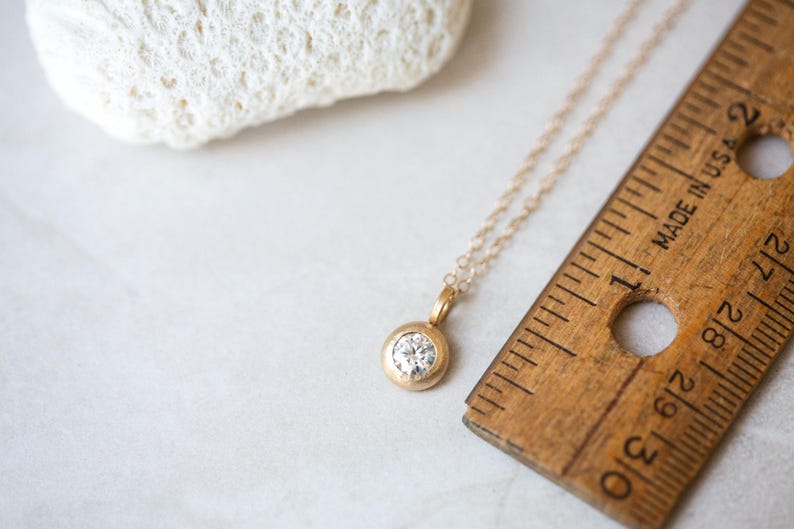 May include: A delicate gold chain necklace with a small round pendant featuring a clear gemstone. The necklace is lying on a white surface next to a wooden ruler.