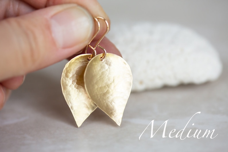 May include: Gold leaf earrings with a hammered texture. The earrings are teardrop shaped and have a medium size.