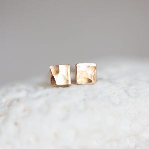 May include: A pair of small, square gold-colored stud earrings. The earrings have a textured surface and are displayed on a white, textured surface. The background is a soft, neutral gray.