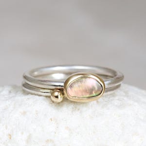May include: A stack of two silver rings with a gold band and a pearlescent stone. A small gold bead sits next to the main ring. The rings are displayed on a white, textured surface.