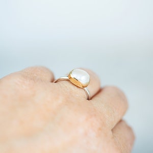 May include: A silver ring with a white oval gemstone set in a gold bezel. The ring is worn on a person's finger.