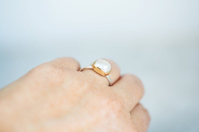 May include: A silver ring with a white oval gemstone set in a gold bezel. The ring is worn on a person's finger.