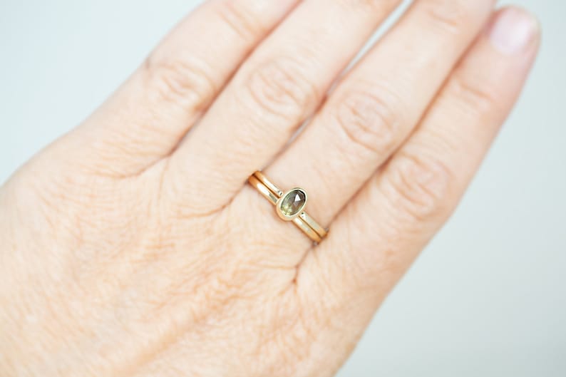 May include: A gold ring with a small, oval-shaped gemstone set in the center. The ring is worn on a finger.