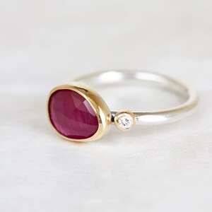 May include: A silver ring with a red ruby gemstone set in a gold bezel. A small diamond is set in the band.