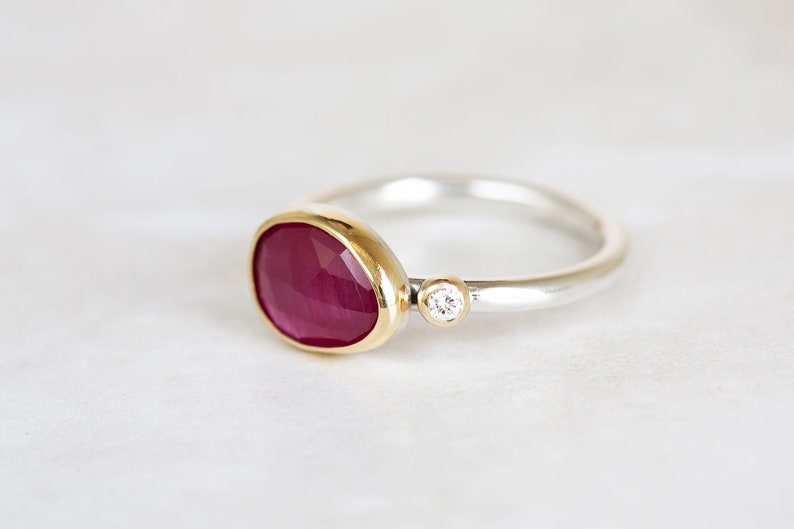 May include: A silver ring with a red ruby gemstone set in a gold bezel. A small diamond is set in the band.