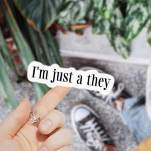 May include: A silver ring with a leaf design is worn on a finger, holding a white sticker that reads "I'm just a they" in black text. The background includes green plants and a person wearing jeans and sneakers.