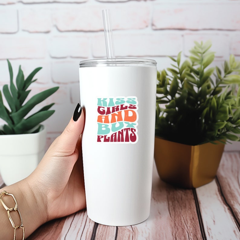Op de afbeelding: Een witte beker met een kleurrijke sticker waarop "Kiss Girls and Buy Plants" staat.