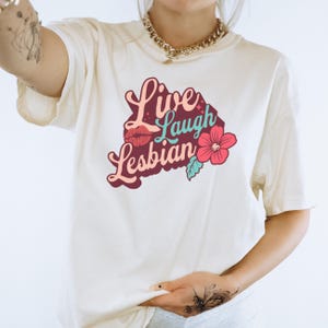 May include: White t-shirt with a maroon graphic that says "Live Laugh Lesbian" with a pink flower and a red lip print.