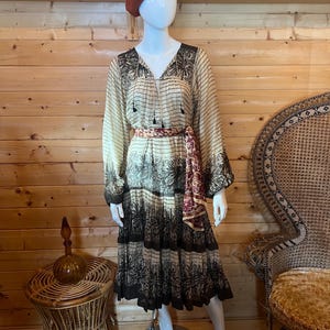 Könnte beinhalten: Ein beigefarbenes und braunes gemustertes Kleid mit langen Ärmeln und einem Stufenrock wird auf einer Schaufensterpuppe präsentiert. Das Kleid ist mit einem gemusterten Gürtel in der Taille gerafft. Auf dem Kopf der Schaufensterpuppe befindet sich ein weinrotes Barett. Ein Beistelltisch und ein Stuhl aus Korbgeflecht befinden sich im Hintergrund.