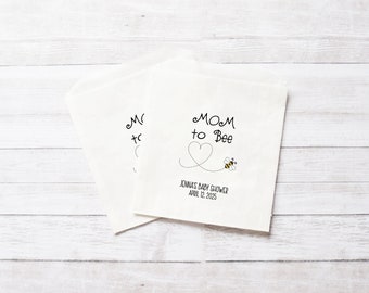 May include: White paper favor bags with black text that reads "Mom to Bee" with a heart and a bee illustration. The text also includes "Jenna's Baby Shower April 12, 2023".