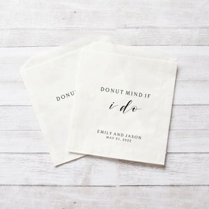 May include: Two white paper bags with the text "Donut mind if I do" in black script font. The bags are for wedding favors and have the names Emily and Jason and the date May 21, 2022 printed on them.