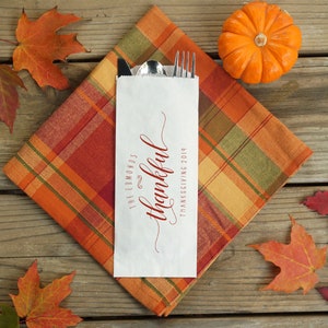 May include: A white paper bag with a red and green plaid napkin, a fork, a spoon, and a knife. The bag has the text "The Edmonds" and "thankful" in cursive script, with the date "Thanksgiving 2019" below. A small orange pumpkin and fall leaves are on a wooden surface.