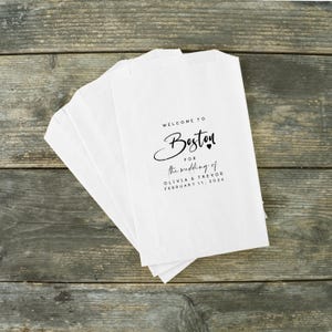 May include: White paper bags with the text "WELCOME TO Boston for the wedding of Olivia & Trevor February 11, 2026" printed in black script. The bags are stacked on a weathered wooden surface.