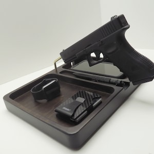 May include: A dark brown wooden valet tray with a black handgun, smartphone, pen, smartwatch, and a carbon fibre wallet. The tray has multiple compartments and a brass support.