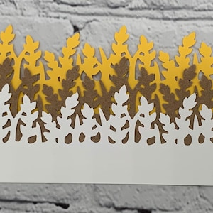 May include: A layered paper craft featuring a repeating leaf design. The top layer is yellow, the middle layer is brown, and the bottom layer is white. The design is cut out, creating a decorative border. The background is a white brick wall.